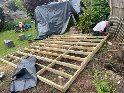 A person constructing a timber deck in a garden surrounded by plants and tools.